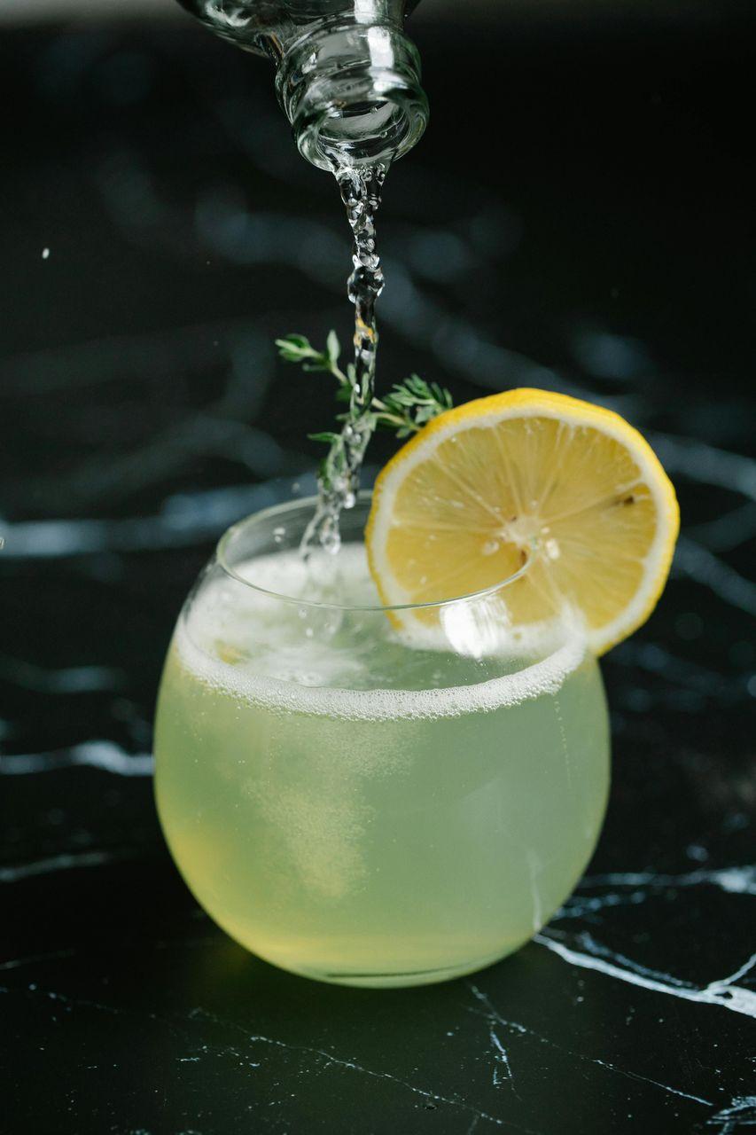 Ein erfrischender gelber Cocktail in einem runden Glas, verziert mit einer Zitronenscheibe und einem Thymianzweig, während sprudelndes Wasser eingeschenkt wird.