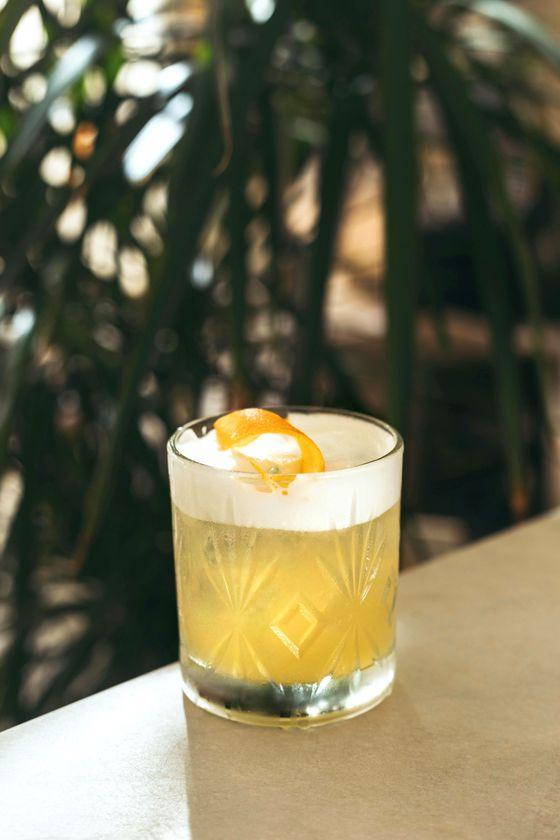 Ein erfrischender gelber Cocktail in einem verzierten Glas mit einer Schaumkrone und einer Orangenschale als Garnitur, serviert bei einem Afterwork-Cocktailabend auf der Dachterrasse.